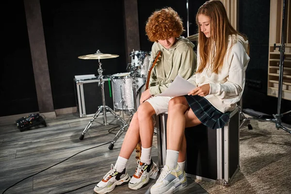 Elegante ragazza adolescente guardando i testi accanto a rosso dai capelli teen tenendo la chitarra in studio di musica — Foto stock