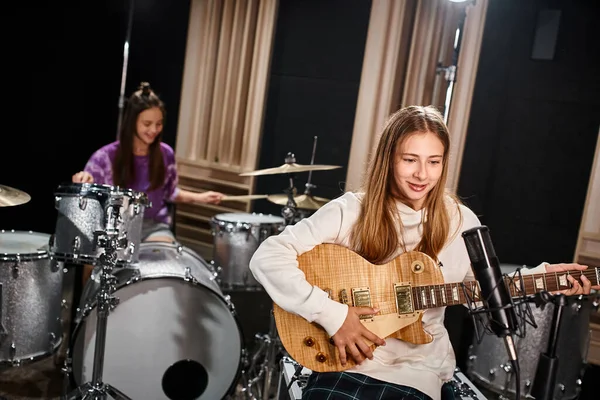 Fokus auf hübsche blonde Teenager-Gitarristin, die neben ihrem verschwommenen Freund spielt, der im Studio Schlagzeug spielt — Stockfoto