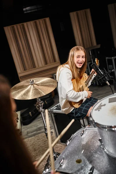 Jolly ragazza adolescente in abito casual suonare la chitarra e guardando il suo amico suonare la batteria — Foto stock