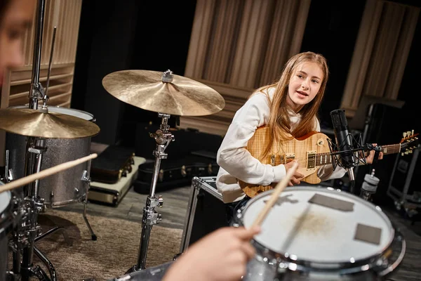 Entzückendes blondes Teenager-Mädchen in lässiger Kleidung, das Gitarre spielt und ihren Freund beim Schlagzeugspielen ansieht — Stockfoto