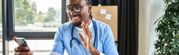Веселий афроамериканський лікар в окулярах махає на камеру телефону і радісно посміхається, банер — стокове фото
