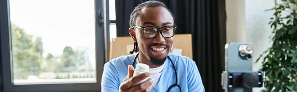 Усміхнений афроамериканський лікар, який показує таблетки на камеру мобільного телефону, телемедицина, банер — стокове фото