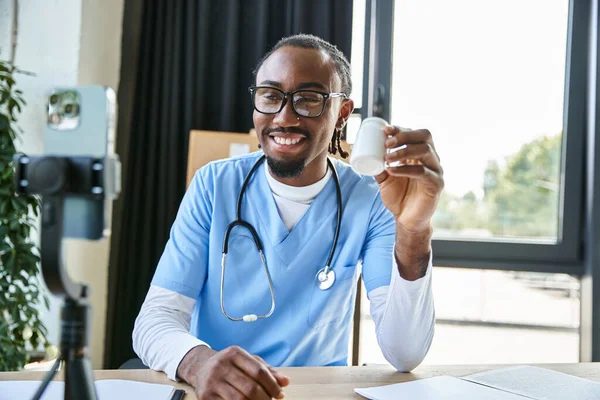 Médecin afro-américain joyeux avec des lunettes montrant des pilules à la caméra de téléphone mobile, télémédecine — Photo de stock
