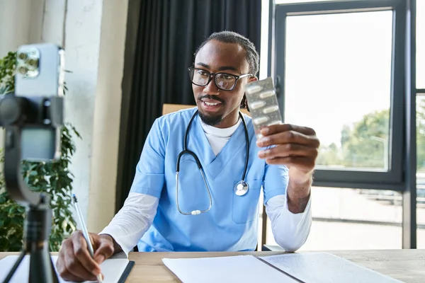 Alegre atraente Africano americano médico segurando pílulas e tomar notas enquanto consulta por telefone — Fotografia de Stock