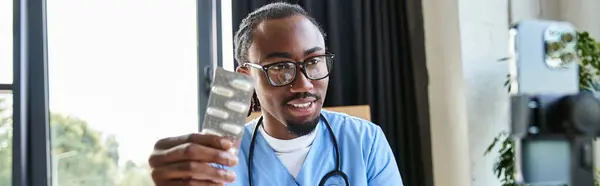Alegre médico afroamericano sosteniendo píldoras y tomando notas mientras consulta por teléfono, pancarta - foto de stock