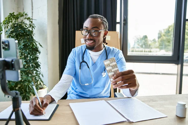 Веселий привабливий афроамериканський лікар тримає таблетки і робить нотатки під час консультації по телефону — стокове фото