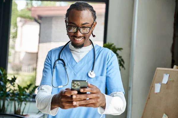 Guapo médico afroamericano feliz con estetoscopio mirando su teléfono móvil, telemedicina - foto de stock