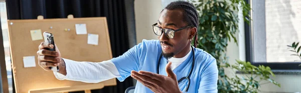 Зосереджений афроамериканський лікар в окулярах консультування пацієнта мобільним телефоном, телевізійним здоров'ям, банером — стокове фото