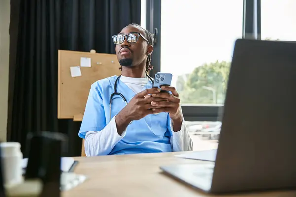 Задумчивый молодой африканский американский врач со стетоскопом, держащий мобильный телефон и отворачивающийся — стоковое фото