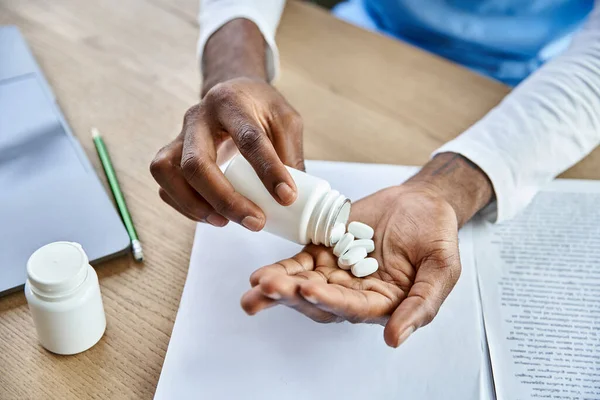 Обрізаний вид на молодого афроамериканського професійного лікаря, який вливає таблетки в руки в його кабінет — стокове фото