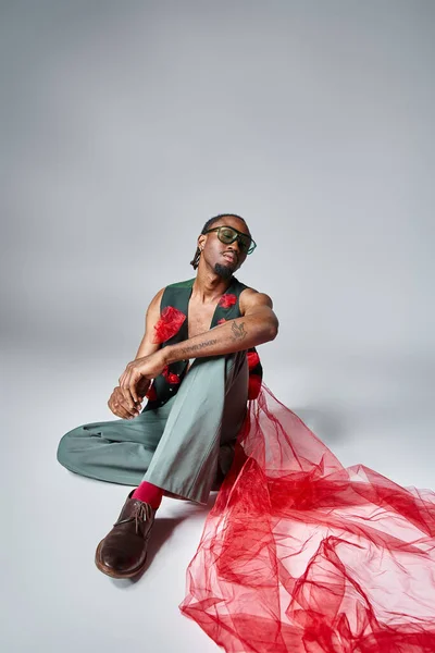 Attrayant homme afro-américain avec du tissu de tulle rouge sur son gilet assis sur le sol, concept de mode — Photo de stock