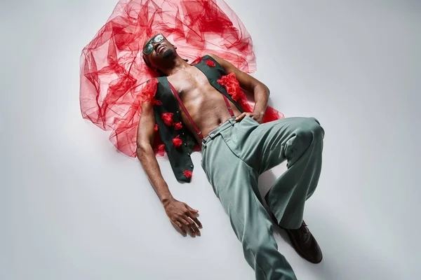Guapo joven afroamericano modelo masculino en traje vibrante con tela de tul rojo acostado en el suelo - foto de stock