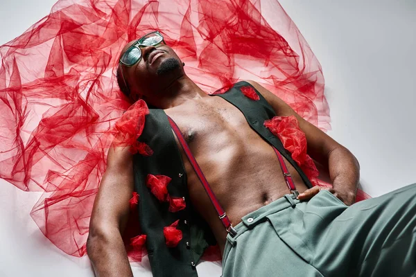 Good looking african american male model in vibrant attire with red tulle fabric lying on floor — Stock Photo