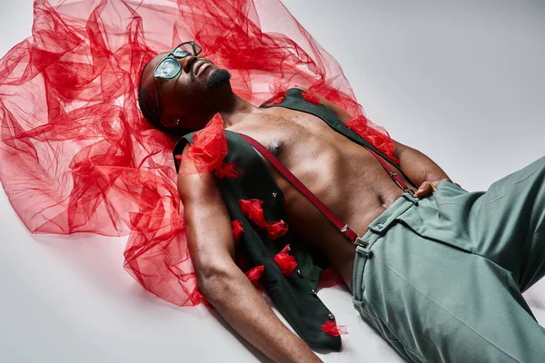 Attractive young african american male model in stylish attire with red tulle fabric lying on floor — Stock Photo