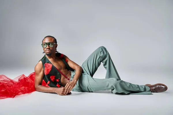 Atractivo hombre afroamericano en traje vibrante con tela de tul rojo reclinado en el suelo, la moda - foto de stock