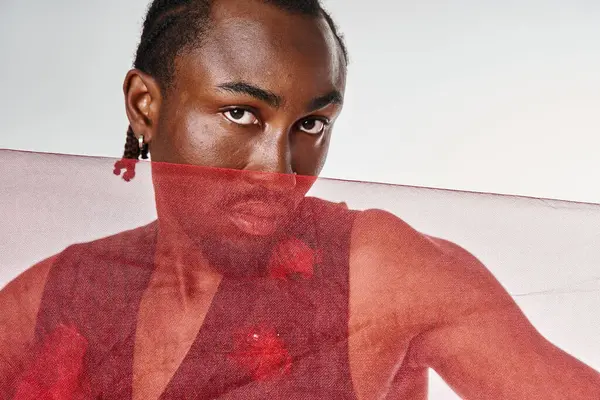 Appealing young african american male model posing with vibrant red tulle fabric on gray backdrop — Stock Photo