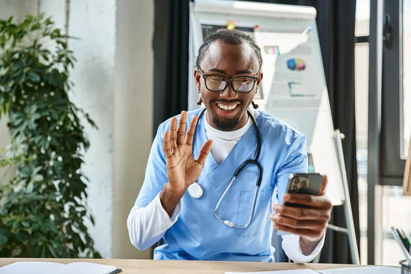 Веселий афроамериканський лікар в окулярах махає на камеру мобільного телефону і щасливо посміхається — стокове фото