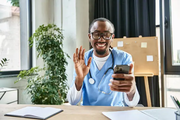 Feliz afroamericano médico con gafas ondeando a la cámara del teléfono móvil y sonriendo felizmente - foto de stock