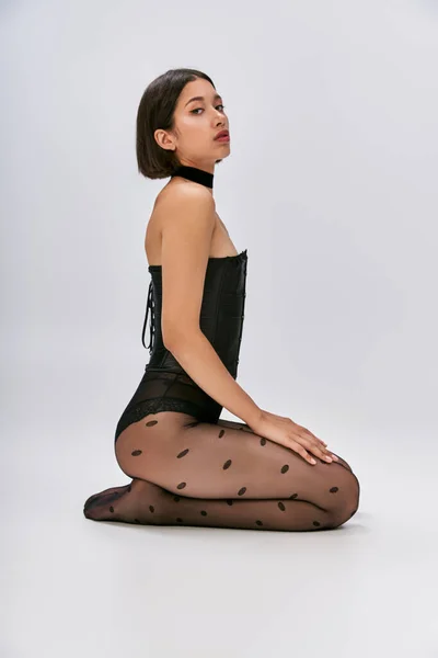 Pretty asian woman with short hair sitting in black corset and polka dot tights on white backdrop — Stock Photo
