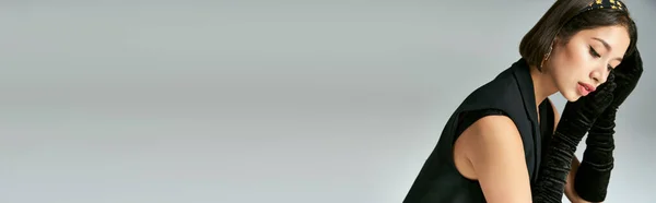 Attraente e sognante donna asiatica in elegante abbigliamento con guanti posa su sfondo grigio, banner — Foto stock