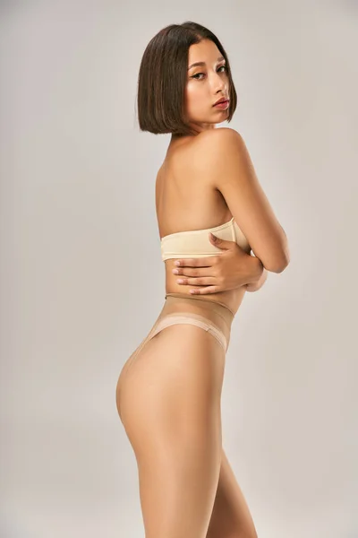 Attractive and young asian woman in seamless pantyhose and beige underwear posing on grey background — Stock Photo