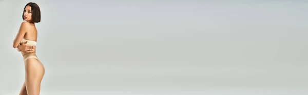 Банер молодої азіатки в безшовних колготках і бежевій білизні позує на сірому фоні — стокове фото