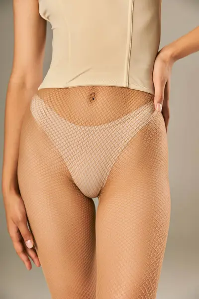 Cropped view of young woman in fishnet tight and panties posing with hand on hip on grey background — Stock Photo