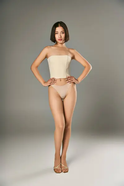 Joven mujer asiática en medias de rejilla y bragas posando con las manos en las caderas sobre fondo gris - foto de stock