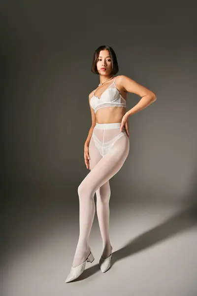 Verführerische asiatische Frau in weißem Spitzen-BH und Strumpfhose posiert mit der Hand an der Hüfte auf grauem Hintergrund — Stockfoto