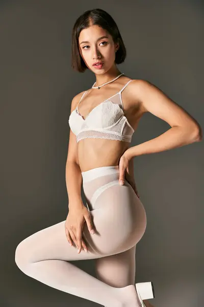 Schöne asiatische Frau in weißer Spitze BH und Strumpfhose posiert mit der Hand auf der Hüfte auf grauem Hintergrund — Stockfoto