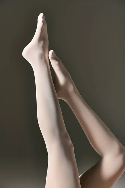 Cropped view of young woman in sheer white pantyhose posing with raised legs on dark grey background — Stock Photo