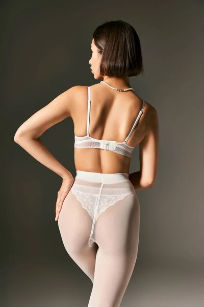 Back view of young woman in white underwear and pantyhose posing with hand on hip on grey background — Stock Photo