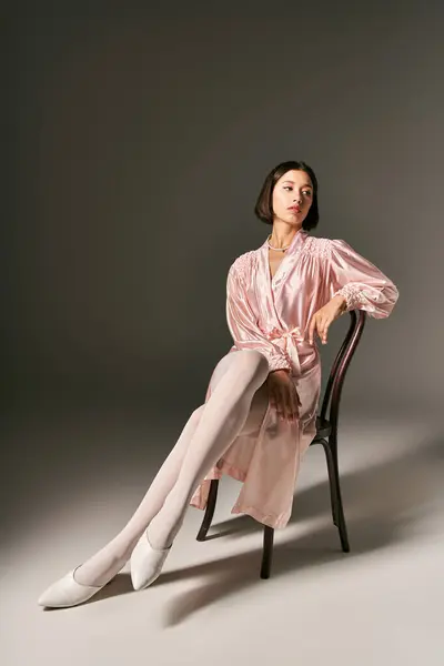 Приваблива молода азіатська жінка в рожевому шовковому халаті та білих колготках, що сидять на стільці на сірому фоні — стокове фото