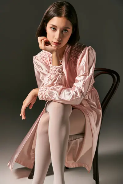 Bonita joven asiática mujer en rosa túnica de seda y medias blancas sentado en silla sobre fondo gris - foto de stock