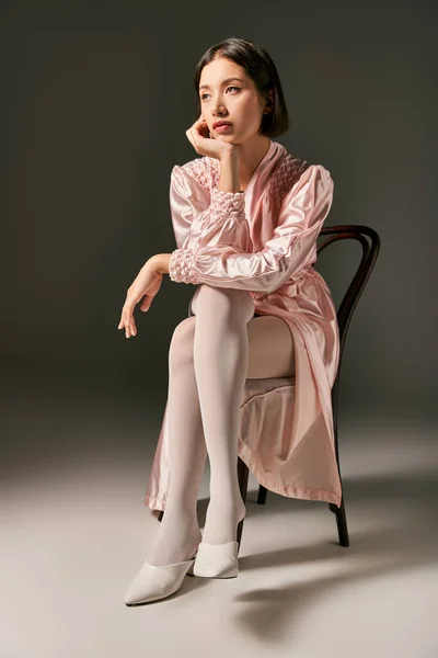 Mujer asiática joven soñadora en túnica de seda rosa y medias blancas sentadas en silla sobre fondo gris - foto de stock