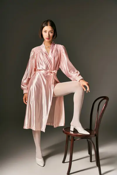 Bonita joven asiática mujer en rosa túnica de seda y medias blancas posando cerca de silla sobre fondo gris - foto de stock