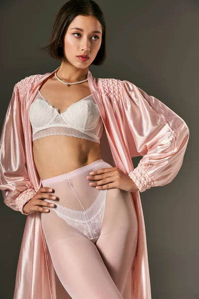 Seducente asiatica giovane donna in abito di seta rosa, reggiseno bianco e collant in posa con le mani sui fianchi su grigio — Foto stock