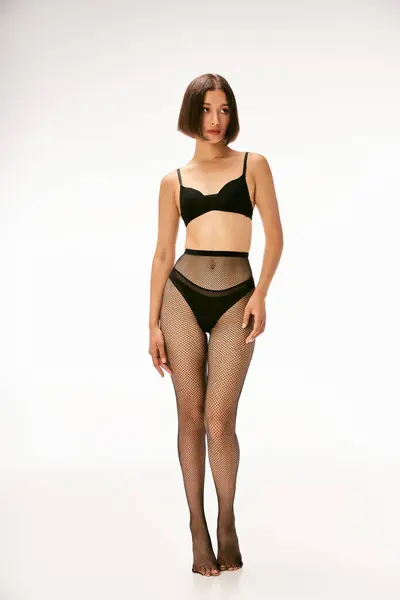 Seducente donna asiatica in calze a rete nere e biancheria intima in piedi su sfondo bianco, calzetteria — Foto stock
