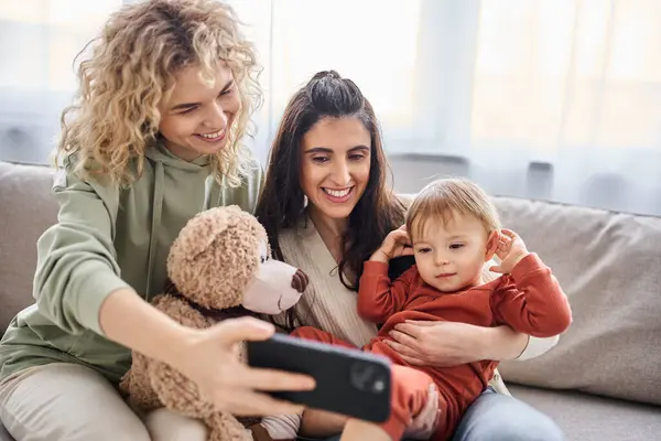 Loving lgbt couple on sofa with their baby girl looking at phone holding teddy bear, family concept — Stock Photo