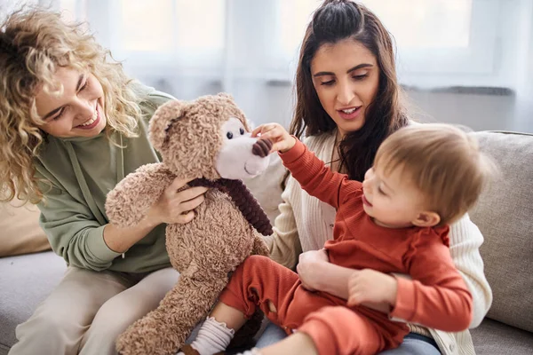 Fröhliches lesbisches Paar mit Kleinkind-Tochter und Teddybär auf Sofa, Familienkonzept — Stockfoto