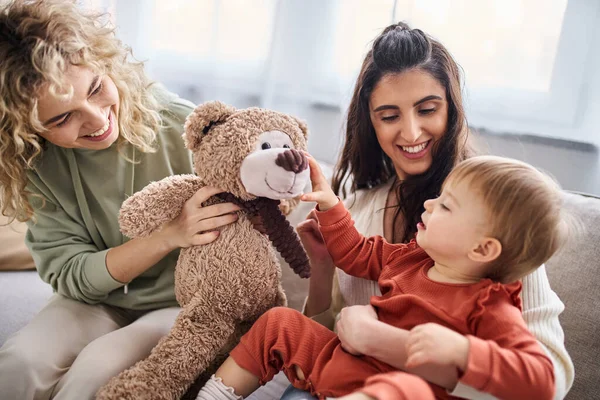 Hermosa pareja lesbiana feliz con su hija pequeña y oso de peluche en el sofá, concepto de familia - foto de stock