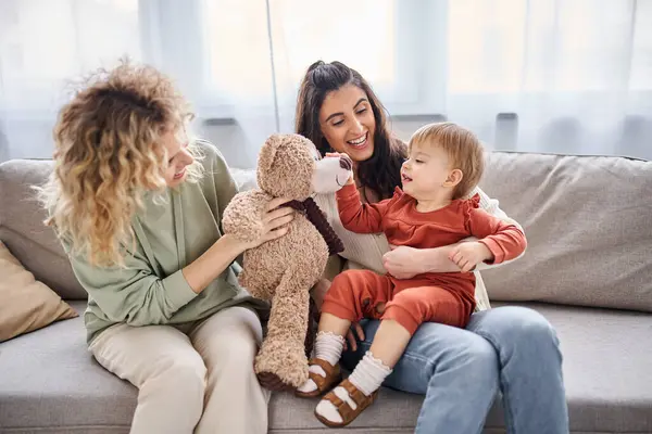 Atractiva pareja lésbica alegre con su hija pequeña y osito de peluche en el sofá, concepto de familia - foto de stock