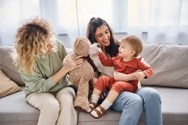 Hermosa pareja lésbica alegre con su hija pequeña y oso de peluche en el sofá, concepto de familia - foto de stock
