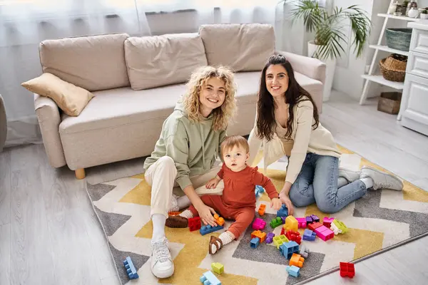 Feliz pareja de lesbianas amorosas jugando con juguetes con su hija pequeña en casa, crianza moderna - foto de stock