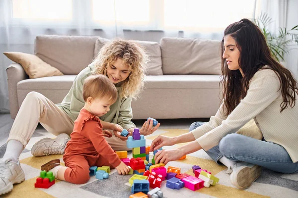 Alegre pareja lesbiana jugando con su bebé niña con juguetes en el suelo en casa, concepto de familia - foto de stock