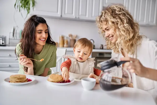 Alegre atractivo lgbt pareja teniendo delicioso desayuno con su bebé niña, concepto de familia - foto de stock
