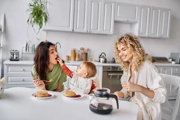 Alegre atractivo lgbt pareja teniendo delicioso desayuno con su bebé niña, concepto de familia - foto de stock