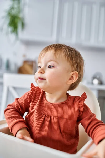 Encantadora niña en suéter naranja sentada en la silla alta en el desayuno y mirando hacia otro lado - foto de stock
