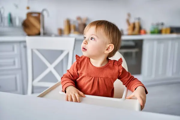 Adorable linda niña en suéter naranja sentado en la silla alta en el desayuno y mirando hacia otro lado - foto de stock