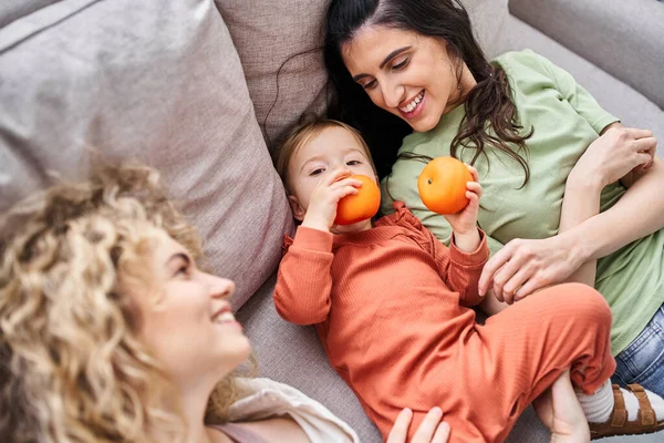 Joyeux couple lgbt couché sur le canapé avec leur jolie fille tenant des mandarines, concept de famille — Photo de stock